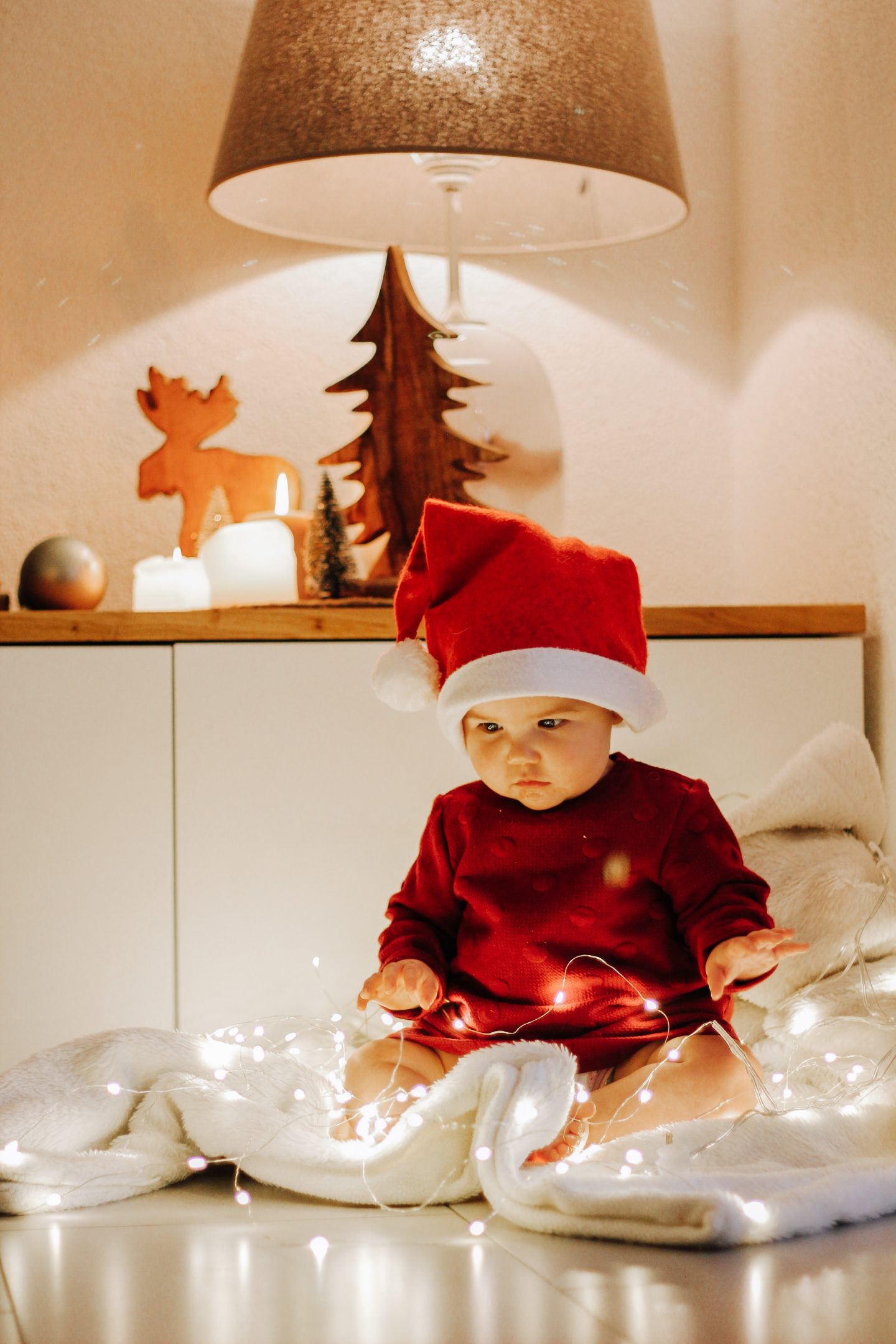 A baby wearing a Santa hat sitting on a cozy blanket looking at a string of beautiful Christmas lights laid out in front of them.